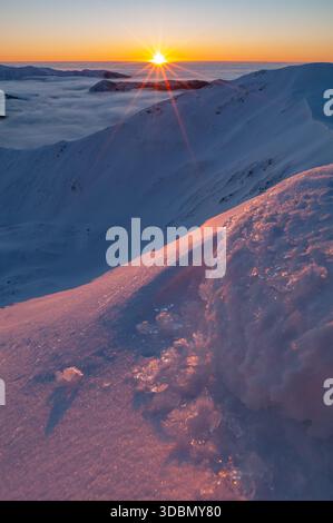 Blick auf die schneereiche Landschaft geküsst von den warmen Farben der aufgehenden Sonne über einem Wolkenmeer, Kotliska, Banska Bystrica Region, Slowakei. Stockfoto