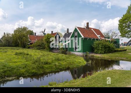 Blick auf lebendige grüne Holzhäuser mit roten Dächern reflektieren das ruhige Wasser eines Kanals unter einem teilweise bewölkten Himmel, Zaandijk Zaanse Schans, Zaanda Stockfoto