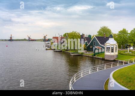 Blick auf traditionelle dunkle Gewächshäuser und rote Windmühlen säumen einen ruhigen Kanal unter einem ruhigen Himmel, Zaandijk Zaanse Schans, Zaandam, Niederlande. Stockfoto