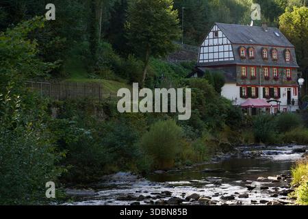 Fachwerkhaus mit Schieferdach und roten Rollläden steht neben einem Bach, umgeben von Grün in Monschau, Nordrhein-Westfalen Stockfoto