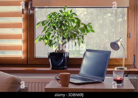 Arbeitsplatz zu Hause mit Laptop. Gemütliches Zuhause im warmen Fenster, das durch Jalousien beleuchtet wird. Stockfoto