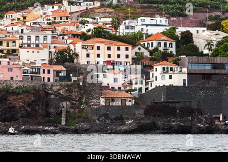 Madeira, pulsierende Küstenlandschaft aus pastellfarbenen und weißen Häusern mit roten Dächern, die entlang zerklüfteter Klippen am Meer thronen, mit Steinmauern und Terra Stockfoto