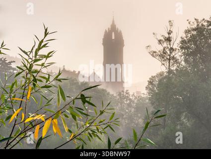 Der alte Kirchturm erhebt sich aus dem Nebel über einer ruhigen Stadt, umgeben von Bäumen und Nebel. Stockfoto