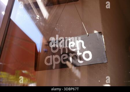 Zweisprachiges Türschild hinter der Fensterscheibe der Eingangstür eines Cafés in Regensburg. Die Sonne fällt durch die Tür auf das Schild. Auf dem Schild steht 'geöffnet' in zwei Sprachen, 'offen' auf Deutsch und 'aperto' auf Italienisch Stockfoto