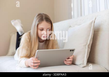 Mädchen, das auf dem Sofa liegt und ein Tablet benutzt. Blondes Mädchen, das sich auf einer weichen Couch entspannt, während es sich auf ein Tablet konzentriert, in einem ruhigen Zuhause stöbert oder studiert Stockfoto