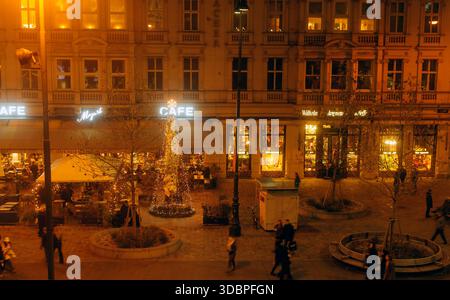 Abendliche Straßenszene mit Leuten, die an beleuchteten Cafés und einem geschmückten Weihnachtsbaum in festlicher urbaner Umgebung vorbeilaufen Stockfoto