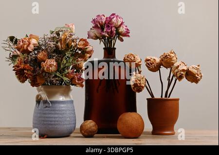 Ein ruhiges Stillleben mit getrockneten Blumenarrangements in einer Mischung aus Keramik, Terrakotta und dunklen Glasgefäßen auf einem Holztisch. Stummgeschaltetes Ohr Stockfoto