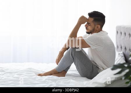 Porträt eines traurigen jungen Mannes aus dem Nahen Osten, der auf einem Bett in einem hellen Schlafzimmer sitzt Stockfoto