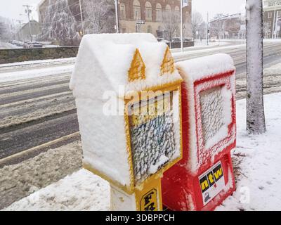Zwei Zeitungskisten sind mit Schnee bedeckt. Eine ist gelb und die andere rot. Der Schnee stapelt sich auf den Postfächern Stockfoto