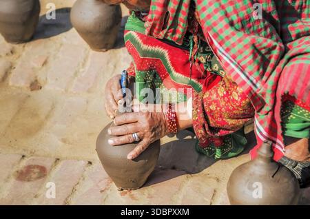 Handwerker in bunten Kleidern und roten Armreifen schmücken Tontopf von Hand, der zwischen Töpferwaren auf sonnendurchfluteten Ziegeln sitzt Stockfoto