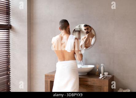 Selbstbewusster junger arabischer Mann, der sich im Badezimmer aufhält und seine Reflexion im Spiegel genießt Stockfoto