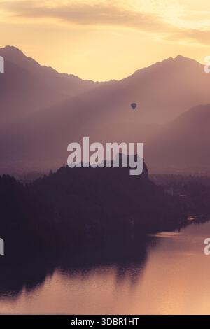 Ein Heißluftballon schwingt über der Silhouette von Bled Castle. Die aufgehende Sonne taucht die Szene in warmes, oranges Licht. Stockfoto