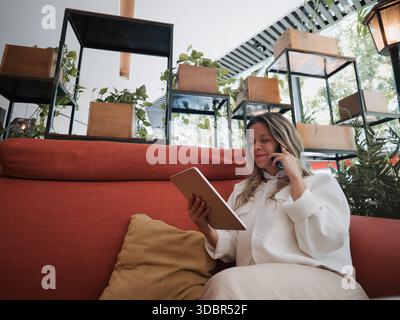 Frau telefoniert, während sie ein Tablet auf einem Café-Sofa benutzt. Stockfoto