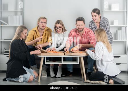 Eine Gruppe von Freunden isst Pizza und lacht zusammen. Fröhliche Freunde genießen Pizza in einem gemütlichen Zimmer. Freundschaft und Lachen machen dieses Essen zu etwas Besonderem Stockfoto
