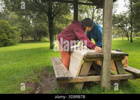 Zwei Frauen arrangieren eine Tischdecke auf einem rustikalen hölzernen Picknicktisch unter einem Unterschlupf Stockfoto