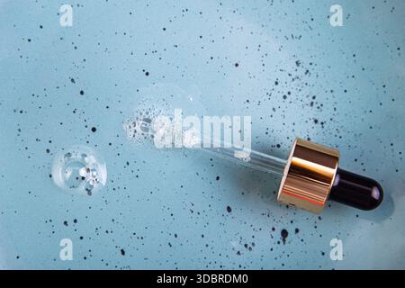 Verschüttetes klares Serum mit Glaspipette auf hellblauem Hintergrund. Flüssiges kosmetisches transparentes Produkt mit kleinen Luftblasen. Flach. Stockfoto