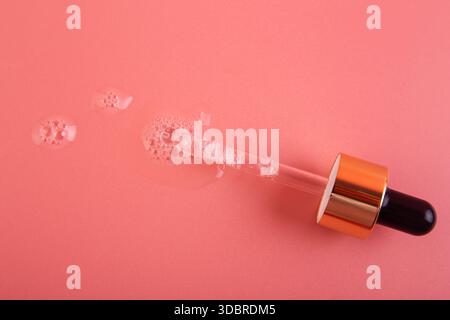 Verschüttetes klares Serum mit Glaspipette auf hellrosa Oberfläche. Flüssiges kosmetisches transparentes Produkt mit kleinen Luftblasen. Flach. Stockfoto