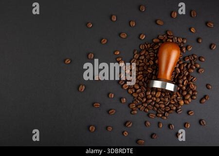 Barista Kit. Kaffeetapfen mit braunem Holzgriff. Draufsicht auf schwarzem Hintergrund mit Kaffeebohnen. Barkeeper-Werkzeug zum Herunterdrücken von gemahlenem Kaffee, A ta Stockfoto