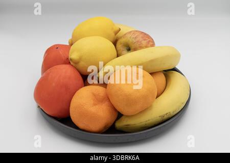 Frische Obstsorten in einer schwarzen Schüssel mit Bananen, Zitronen, Orangen, Äpfeln und Persimmonen isoliert auf weißem Hintergrund Stockfoto