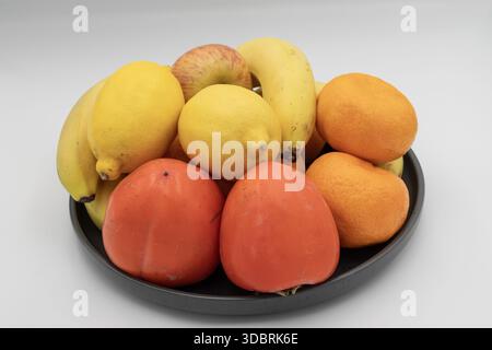 Frische Obstsorten in einer schwarzen Schüssel mit Bananen, Zitronen, Orangen, Äpfeln und Persimmonen isoliert auf weißem Hintergrund Stockfoto