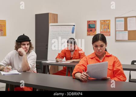 Drei junge Erwachsene Frauen sitzen an Schreibtischen im Klassenzimmer, zwei Frauen in orangefarbenen Gefängnisuniformen lesen und schreiben, eine kaukasische Frau beobachtet, pädagogische Plakate an der Wand Stockfoto