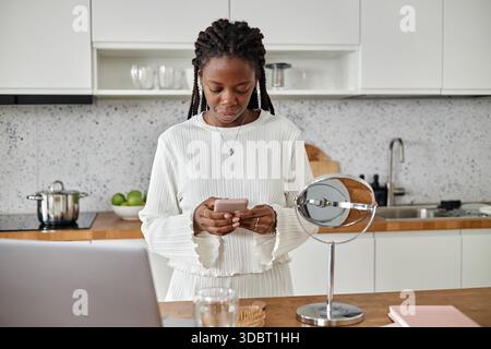 Junge Schwarze Frau, die ihr Smartphone benutzt, während sie in der modernen Küche steht Stockfoto