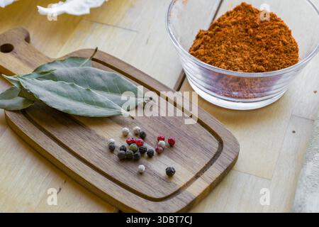 Lorbeerblätter und gemischte Pfefferkörner auf einem hölzernen Schneidebrett mit gemahlenen Gewürzen in einer Glasschale auf einem Küchentisch. Stockfoto