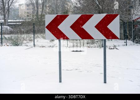 Richtungsschild mit roten und weißen Chevron-Pfeilen auf Metallpfosten, im Freien bei Schneefall fotografiert; schneebedeckter Boden. Stockfoto