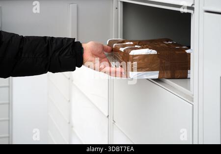 Nahaufnahme einer menschlichen Hand in einem schwarzen Jackenärmel, die ein rechteckiges Paket aus einem grauen Metallfach zieht. Stockfoto