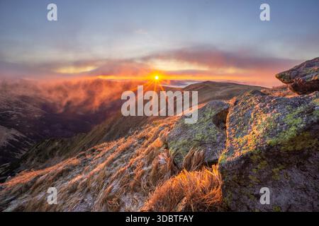 Blick auf die Sonne, die durch die Wolken über felsige Hänge, goldene Gräser und ferne Gipfel durchdringt, Skalka, Banska Bystrica Region, Slowakei. Stockfoto