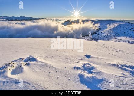 Blick auf einen Sonnenaufgang über einer schneebedeckten Landschaft mit Wolkenschichten zwischen Berggipfeln, Kosarisko, Banska Bystrica Region, Slowakei. Stockfoto
