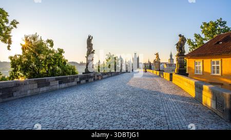 Sonnenlicht beleuchtet die Karlsbrücke in Prag, während der Tag beginnt und sanfte Schatten auf die Kopfsteinpflaster wirft. Die Statuen stehen still, mit Blick auf die friedliche Moldau und den pulsierenden Damm. Stockfoto