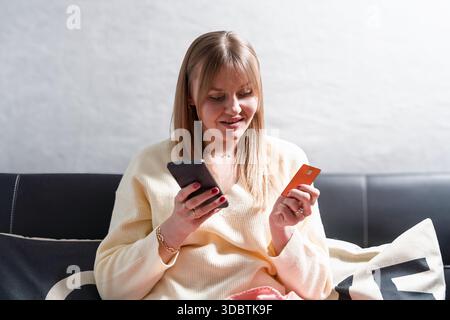Frau, die eine Online-Zahlung mit ihrem Smartphone und einer Zahlungskarte von einem schwarzen Sofa tätigt Stockfoto