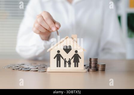 Familienspar- und Finanzplanungskonzept. Eine Frau platziert eine Münze in eine hölzerne, hausförmige Schweinebank mit einem Familiensymbol, das Sparguthaben repräsentiert. Stockfoto