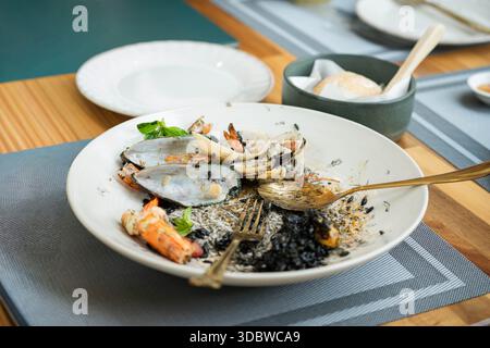 Nahaufnahme eines weißen Tellers mit Resten aus einer schwarzen Tintenschale mit Meeresfrüchten, einschließlich Muschelschalen und Garnelen. Fertig gemahlenes Essen auf einem Holztisch in einer Resta Stockfoto