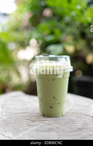 Erfrischende Matcha Latte mit Milchschaum in einer Plastiktasse zum Mitnehmen. Auf einem rustikalen Holztisch mit einem verschwommenen grünen Hintergrund. Ideal für Stockfoto