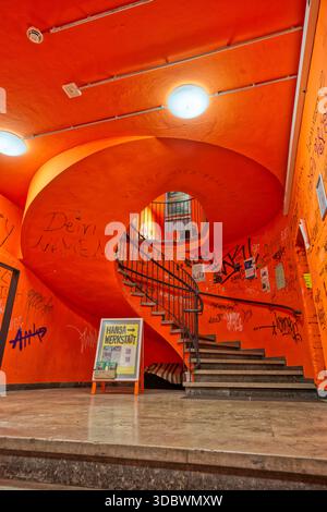 Ein Blick nach oben vom Erdgeschoss auf eine lebendige rote Wendeltreppe in einem städtischen Gebäude. Die Wände sind mit schriftlichen Markierungen bedeckt. Stockfoto