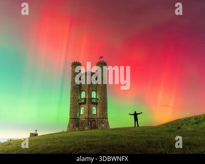 Blick auf eine Silhouette einer Person mit ausgestreckten Armen gegen einen leuchtenden Himmel mit aurora, der in der Nähe eines Steinturms auf einem grasbewachsenen Hügel steht, Broadway to Stockfoto