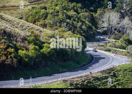 Blick auf eine Serpentinenstraße, die sich durch grüne Hügel schlängelt, wo Sonnenlicht den Asphalt und das üppige Laub in einem lebendigen Tanz aus Licht und Schatten färbt, Tā Stockfoto