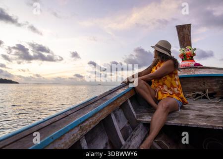 Blick auf eine Frau mit Hut und gelbem Kleid sitzt auf einem Holzboot, während die Sonne über dem ruhigen Meer untergeht und den Himmel in sanften Tönen malt, Phi Phi Island, Kr Stockfoto