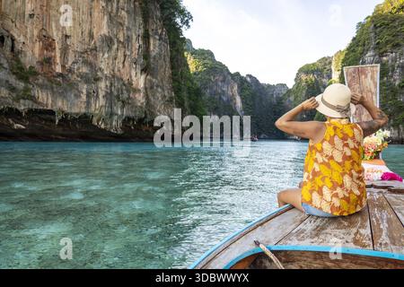 Blick auf eine Frau mit gelbem Dach und Hut auf der Vorderseite eines hölzernen Bootes, das durch türkisfarbenes Wasser mit hohen Klippen segelt, Phi Phi Island Stockfoto