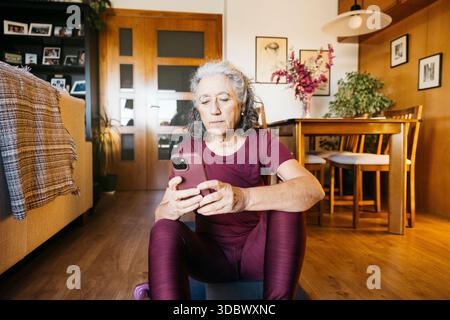 Frau in Sportswear sitzt auf einer Trainingsmatte und überprüft ihr Handy im Wohnzimmer Stockfoto