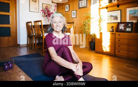 Eine ältere Frau sitzt im Wohnzimmer auf einer Yogamatte und lächelt in die Kamera Stockfoto