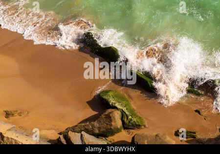 Blick auf schäumende Wellen, die auf moosbedeckte Felsen an einem goldenen Sandstrand prallen, wo das Meer auf die Küste trifft, Gwadar, Belutschistan, Pakistan. Stockfoto