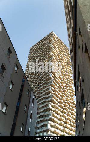 Der Blick auf einen modernen Wolkenkratzer mit einer strukturierten Fassade erhebt sich dramatisch vor einem klaren blauen Himmel, eingerahmt von der starken Geometrie der umliegenden Gebäude, Stockfoto