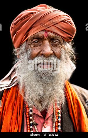 Ein Porträt eines älteren Mannes, möglicherweise eines Sadhu oder Dorflehrten aus Indien, der einen orangefarbenen Turban, einen Schal und einen langen grauen Bart gegen einen schwarzen bac trägt Stockfoto