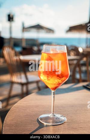 Aperol spritzt Cocktail mit Eis in einem Weinglas auf einem Kaffeetisch am Meer. Erfrischender Sommer-Aperitif mit verschwommenem Strandhintergrund, Nahaufnahme Stockfoto