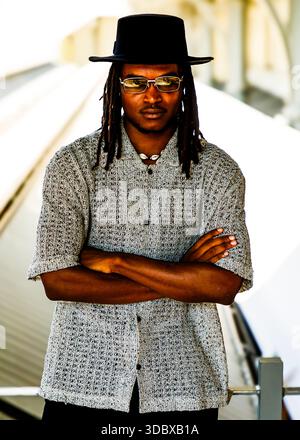 Ein Mann mit Dreadlocks, mit schwarzem Hut und Sonnenbrille, steht mit überkreuzten Armen. Stockfoto
