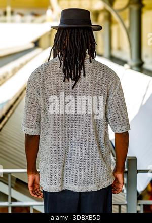 Eine Person mit Dreadlocks, die einen Hut und ein helles Hemd trägt, steht mit dem Rücken zur Kamera. Stockfoto