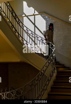 Ein Mann mit Hut steht auf einer geschwungenen Treppe und hält sich am Geländer fest. Die Treppe ist in einem Gebäude mit gekachelten Wänden und einem Schild mit der Aufschrift 'SUBI Stockfoto